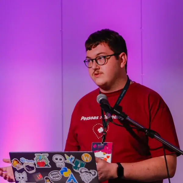 Um jovem de óculos e bigode palestra em um pódio com microfone. Ele usa uma camiseta vermelha e mexe em um notebook coberto de adesivos de tecnologia, com um fundo roxo.